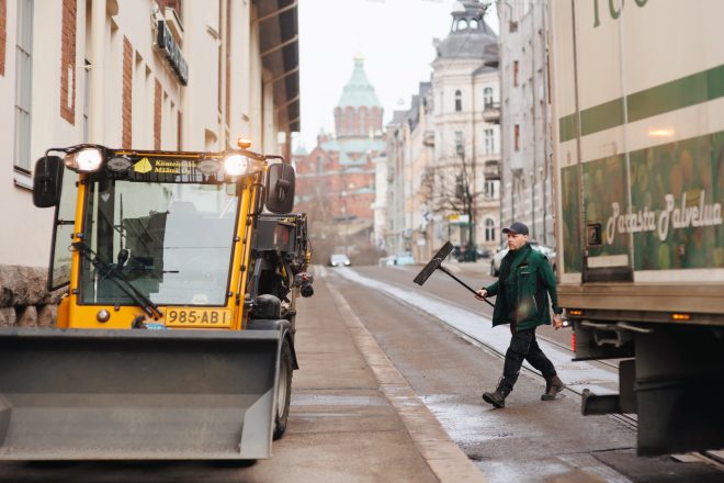 Kaupungin katunäkymä: keltainen aura-auto vasemmalla, oikealle kulkeva mies vihreässä työtakissa kantaa lapioa, taustalla vanhoja kerrostaloja ja kirkontorni.