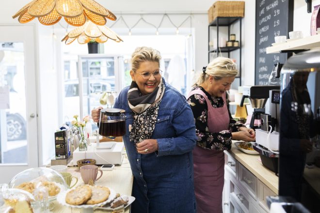 Kahvilan valoisa sisätila, jossa kaksi hymyilevää naista työskentelee tiskin takana — toinen kantaa kahvipannua ja toinen käyttää espressokonetta, etualalla tarjoiluastioita ja leivonnaisia.