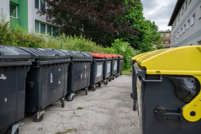 Rivissä suuret pyörälliset jätesäiliöt taloyhtiön pihalla; etualalla keltakansinen säiliö oikealla ja rivissä tummanharmaita säiliöitä punaisilla kansilla, taustalla kerrostaloja ja vihreää puustoa