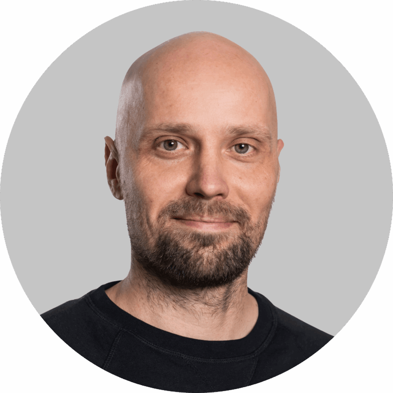 Muotokuva hymyilevästä kaljusta miehestä, jolla on lyhyt parta ja tumma paita, neutraali vaalea tausta.