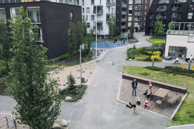 Ilmakuva Helsingin Jätkäsaaren suuresta korttelipihasta, jota ympäröivät modernit asuinkerrostalot; piha-alueella polkuja, nurmikkoa, puita ja pensaita, sininen leikkialue taustalla sekä puinen oleskelulava etualalla, jossa aikuisia ja lapsia istumassa ja leikkimässä.