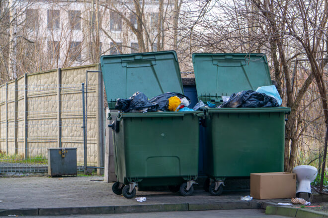 Kaksi suurta vihreää roska-astiaa avoimina ja täynnä roskapusseja, jätteitä levällään astioiden ympärillä, laatikko ja valkoinen sanko maassa, taustalla aita, lehdettömiä puita ja kerrostalo.