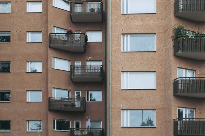 Oranssinkellertävän kerrostalon julkisivu, jossa rivissä ikkunat ja pienet metallikaiteelliset parvekkeet, muutamilla parvekkeilla ruukkuistutuksia ja yhdellä parvekkeella näkyvä henkilö.